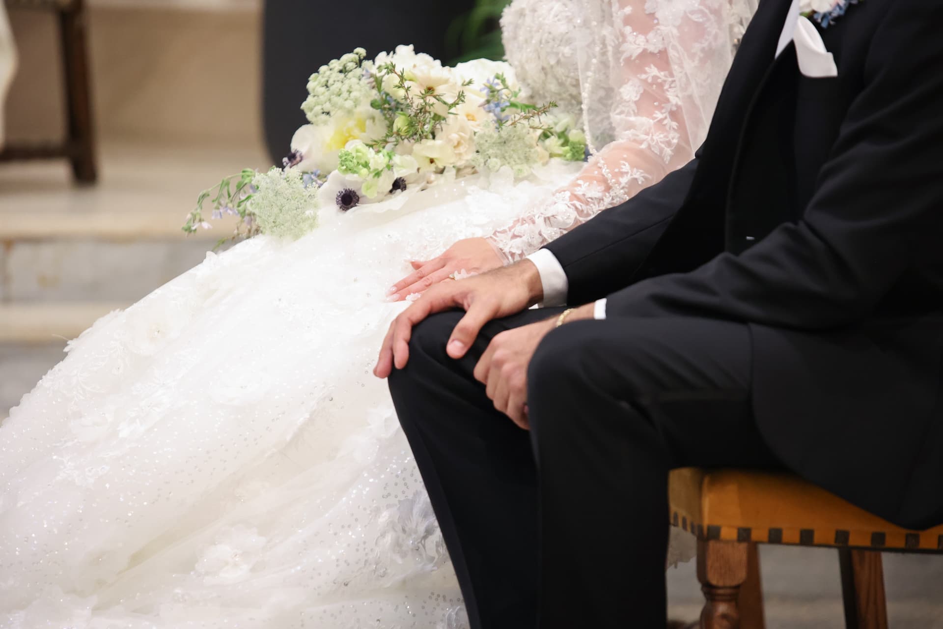 Bride and groom seated side by side during a wedding ceremony, the bride's lace gown and floral bouquet resting in her lap beside the groom's black suit, Austin, Texas