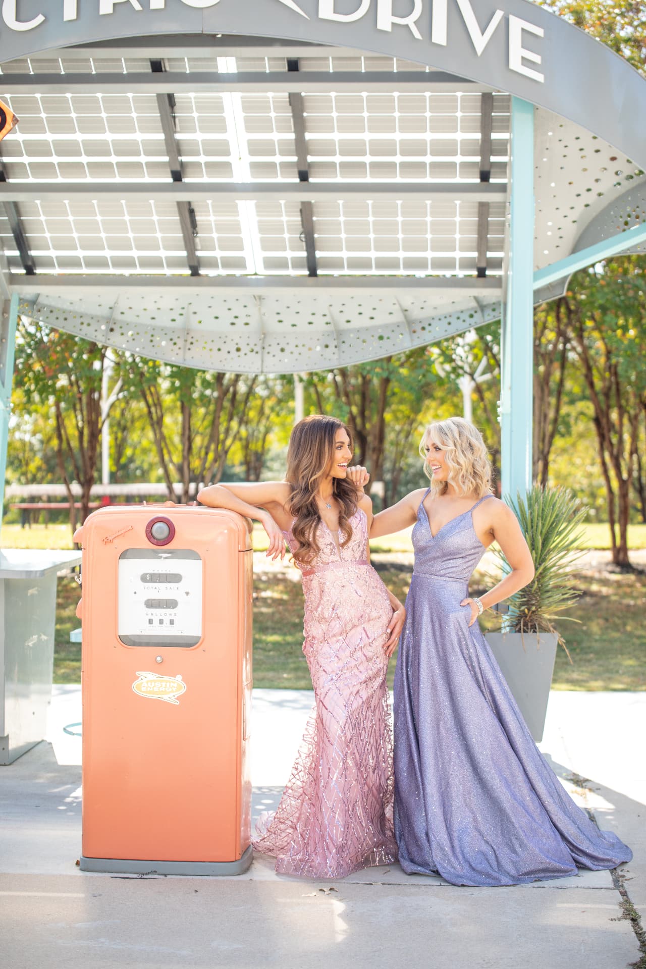 Two young women in sequined formal gowns posing together beside a vintage electric charging station under a solar canopy in Austin, Texas