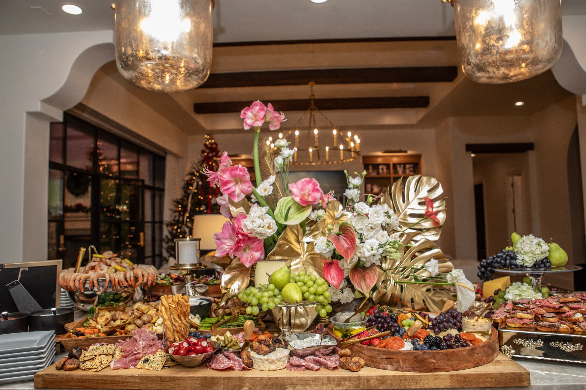 Elegant event buffet spread with a floral centerpiece, charcuterie, fruit and seafood on a wooden table under warm pendant lights at a catered Austin celebration.